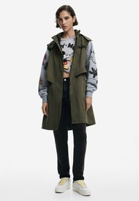 Un trench-coat sans manches vert olive sur un sweat-shirt gris avec des impressions graphiques colorées. Un jean noir et des baskets blanches avec des accents jaunes.