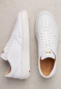 Bílé kožené tenisky s perforovaným designem na špičce, plochými tkaničkami, zlatým logem na jazyku a gumovou podešví s jemnou strukturou.