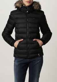 Black puffer jacket with a faux fur-trimmed hood, horizontal quilted design, zip closure, and two side pockets. Accented with logo patch.