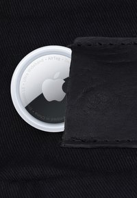 Apple AirTag partially tucked into a black leather pocket on black fabric, showing the Apple logo and circular white edge.