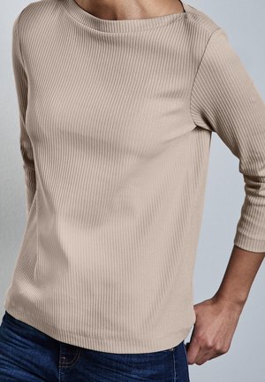 Persoon die een beige geribd shirt met lange mouwen en een donkerblauwe spijkerbroek draagt, staande met één hand op de heup.