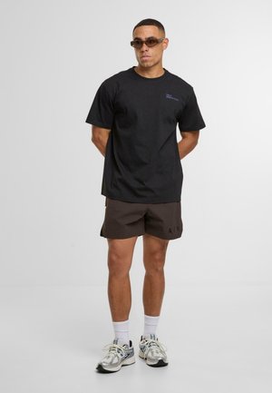 Zwarte katoenen t-shirt met korte mouwen, met een klein blauw logo. Gepaard met bruine shorts en witte crew-sokken, compleet met hardloopschoenen.