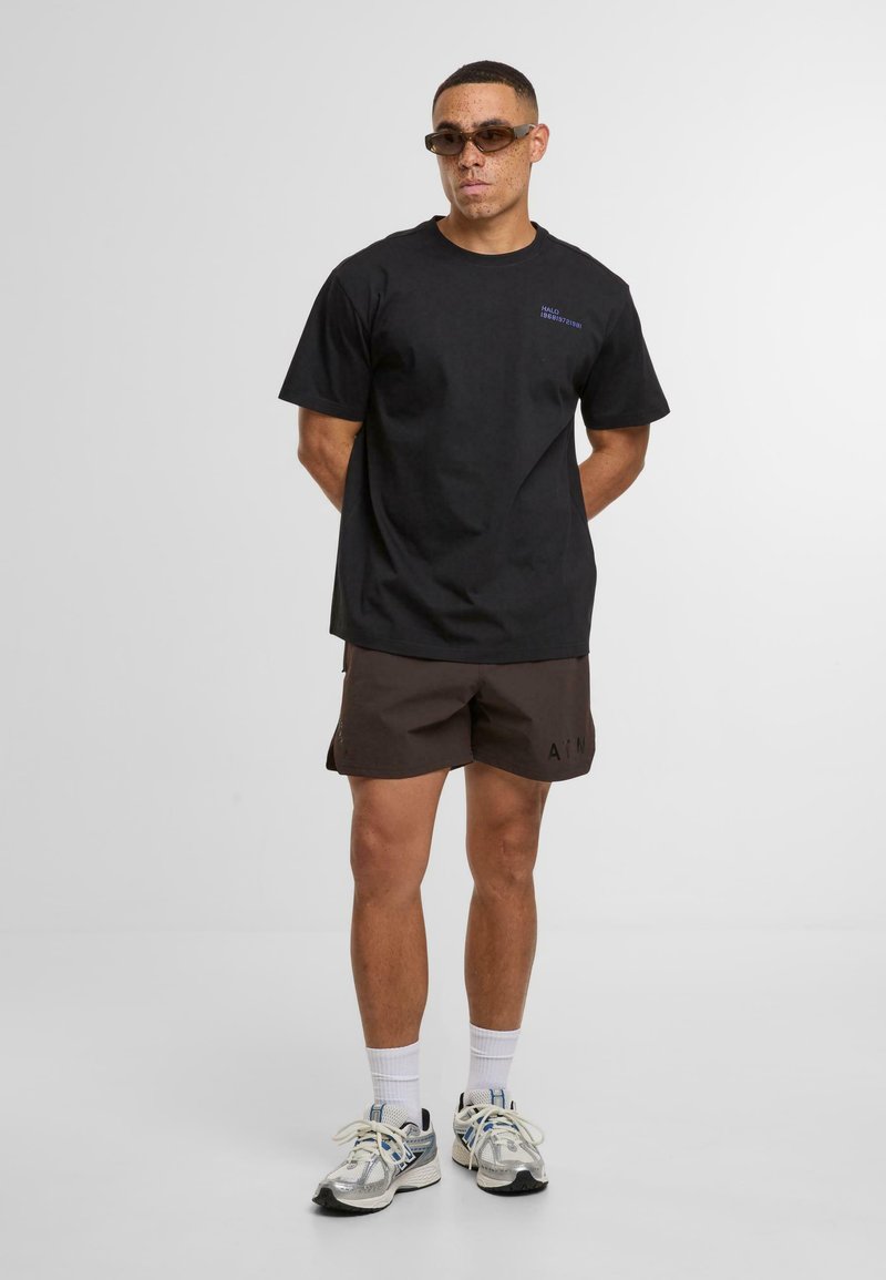 Homme portant un t-shirt noir oversize, un short foncé, des chaussettes blanches, des chaussures de running argentées et des lunettes de soleil, debout devant un fond blanc uni.