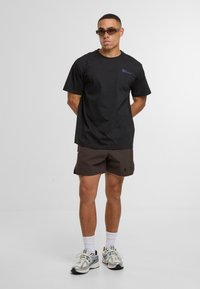 Černé bavlněné tričko s krátkým rukávem, zdobené malým modrým logem. V kombinaci s hnědými šortkami a bílými ponožkami do půli lýtek, doplněné běžeckými teniskami.