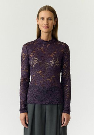 Femme aux cheveux bruns raides portant un haut en dentelle florale violet à manches longues et une jupe gris foncé, debout devant un fond clair uni.