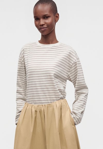 Jeune femme aux cheveux courts portant une chemise blanche à manches longues avec de fines rayures noires et une jupe beige froncée, les mains dans les poches, souriante.