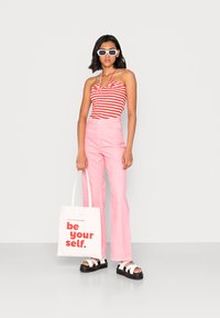 Top sem alças listrado em vermelho e branco, calças flare rosa de cintura alta, óculos de sol brancos e sandálias pretas chunky; segurando uma bolsa de tela.