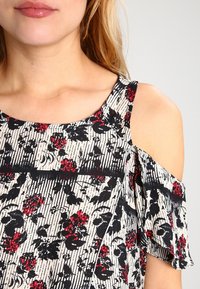 Haut à épaules dénudées en tissu léger, avec un fond blanc à rayures noires et motifs floraux rouges. Manches volantées pour plus de détails.