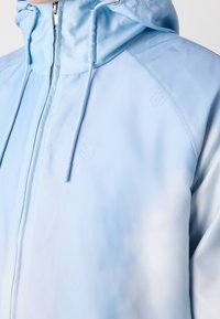 Veste à capuche bleu clair avec fermeture éclair à l'avant et cordons, montrée sur le haut du torse sur un fond uni.