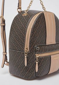 Sac à dos marron à motifs avec des accents crème, des fermetures éclair dorées et une bandoulière chaîne. Comprend une poche avant et une forme arrondie. Surface texturée.