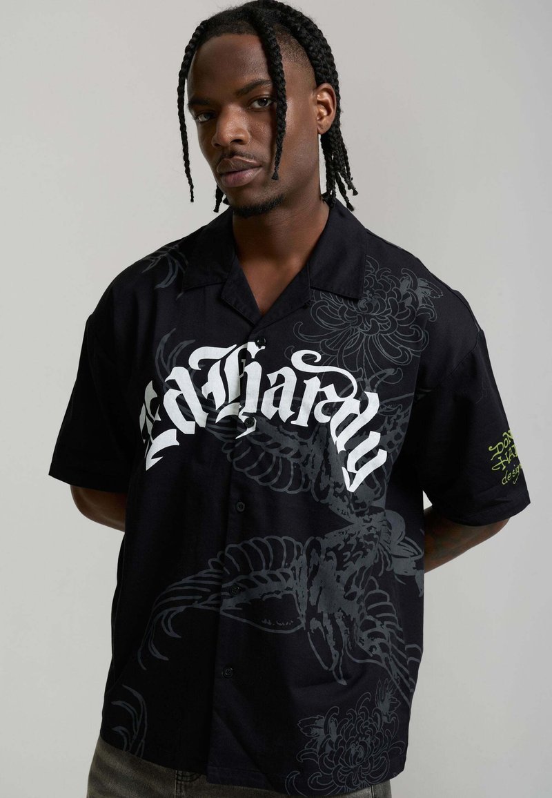 Man with braided hair wearing black button-up shirt with white gothic text and dark floral patterns, standing against plain background.