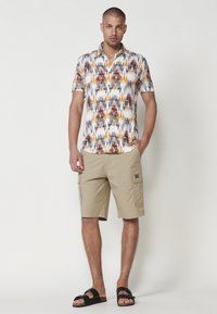 Chemise à manches courtes avec un fond blanc et un motif abstrait multicolore ; associée à un short cargo beige et des sandales noires.