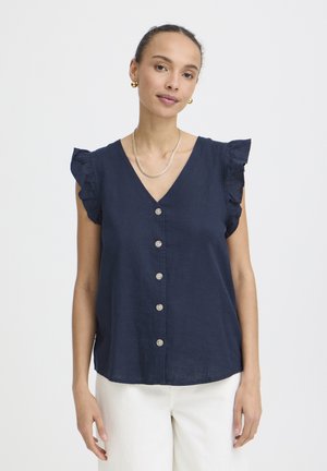 Kobieta ubrana w granatową, bezrękawnikową bluzkę z falbankami na ramionach oraz białe spodnie, uzupełniona złotymi kolczykami i naszyjnikiem.