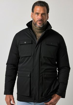Mand med skæg iført sort jakke med fire klaplommer og en høj krave over en brun strikket rullekravesweater.