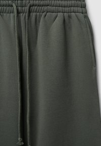 Pantaloni sportivi verde scuro con vita elastica, lacci regolabili, tasca laterale e una consistenza morbida e opaca.