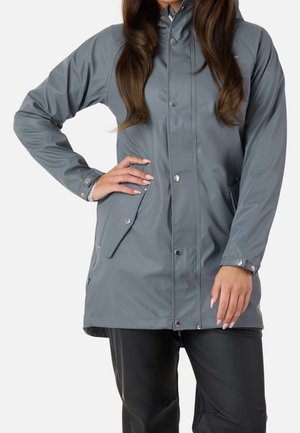 Femme portant une veste imperméable grise avec des poches à boutons et un pantalon noir, debout avec une main sur la hanche devant un fond uni.