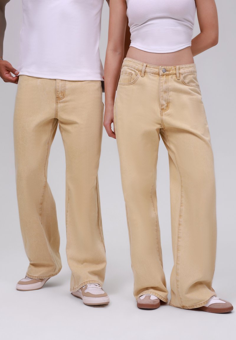 Deux personnes portant des jeans beige à jambes larges, des hauts blancs et des baskets beiges, debout côte à côte sur un fond neutre.
