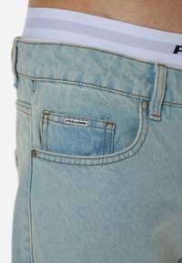 Svetlomodré džínsové šortky s jemnou textúrou, s medeným prešitím, malou zadnou kapsou a bielym pásom so sivými pruhmi.