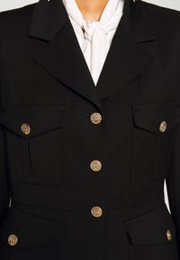 Veste noire taillée avec des boutons dorés texturés et quatre poches à rabat, portée sur une écharpe blanche nouée au cou.