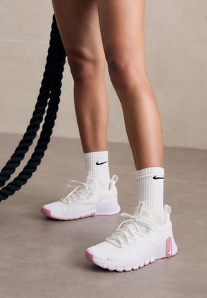 Jambes portant des baskets Nike blanches et roses et des chaussettes Nike blanches, debout sur un sol gris à côté d'une corde de combat noire.
