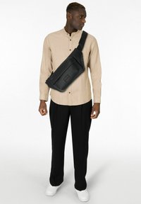 Schwarze Leder-Umhängetasche mit strukturierter Oberfläche, verstellbarem Riemen und vordere Reißverschlusstasche, getragen über einem beigefarbenen Langarmshirt und schwarzen Hosen.