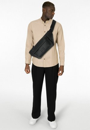 Borsa a tracolla in pelle nera con superficie texturizzata, tracolla regolabile e tasca frontale con zip, indossata sopra una camicia beige a maniche lunghe e pantaloni neri.