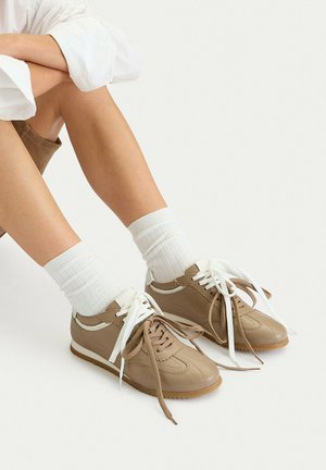 Persona che indossa scarpe da ginnastica color sabbia con lacci multicolore e calzini bianchi, seduta a gambe incrociate su uno sfondo semplice.