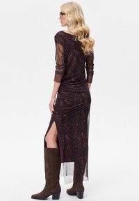 Brązowa sukienka maxi w wzór wężowej skóry z rękawami do połowy przedramienia i rozcięciem z boku, zestawiona z ciemnobrązowymi kozakami do kolan.