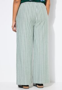 Vaaleanvihreät leveälahkeiset housut valkoisilla pystysuorilla raidoilla, joissa on joustava vyötärönauha ja sileä, teksturoitu kangas. Mustat kengät näkyvissä.