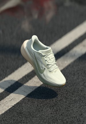 White Nike running shoe with green swoosh mid-air above asphalt with white lines, motion creating small debris and shoe shadow below.