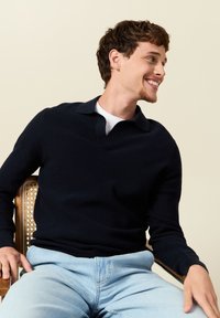 Laivastonsininen neulepusero, jossa on V-pääntie ja kaulustyyli, yhdistettynä vaaleansinisiin farkkuihin. Kankaan pinta on teksturoidun näköinen.