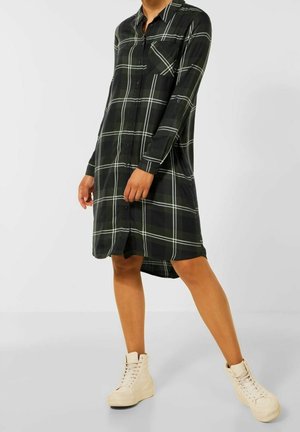 Robe-chemise à carreaux noirs avec manches longues, fermeture à boutons sur le devant et une poche poitrine. Portée avec des baskets montantes crème.