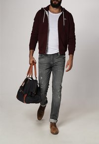Burgundy zip-up hoodie, vit T-shirt, slitna grå jeans, bruna skor. Svart väska med bruna accenter i handen. Avslappnad outfit.