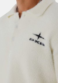 Weißes Fleece-Poloshirt mit strukturierter Oberfläche, kurzem Kragen und schwarzem Logodetail. Verfügt über drei Knöpfe entlang der Knopfleiste.