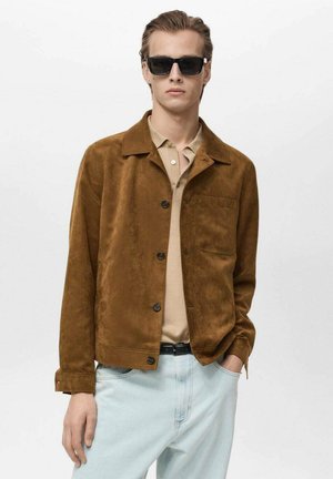 Bruine suède jas met een kraag, knopen aan de voorkant en een borstzak, in combinatie met een beige poloshirt en lichtblauwe spijkerbroek. Zwarte zonnebril gedragen.