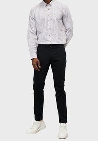 Camicia bianca a maniche lunghe con bottoni, caratterizzata da un motivo sottile, abbinata a pantaloni neri slim-fit e scarpe da ginnastica bianche. Sono visibili dettagli con bottoni.