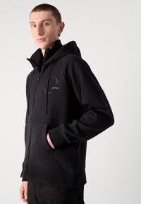 Hoodie zippé noir en matériau doux, avec un col haut, des poignets côtelés et un logo sur la poitrine. Accentué par une poche kangourou.