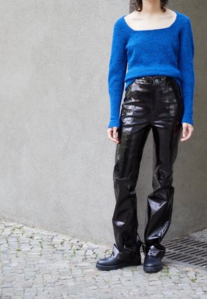 Un pull en maille bleu avec un col carré, associé à un pantalon en vinyle noir brillant et des chaussures noires imposantes sur un surface en pavés.