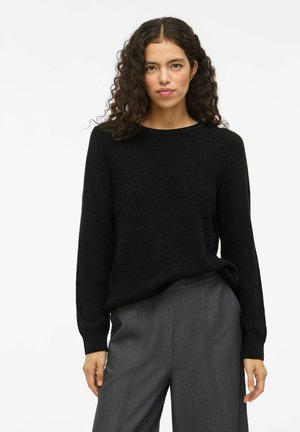 Frau mit lockigem Haar, die einen schwarzen Strickpullover und graue maßgeschneiderte Hosen trägt und vor einem schlichten hellen Hintergrund steht.