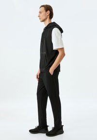 Sudadera sin mangas negra con bolsillo frontal y cordón, combinada con pantalones ajustados negros y zapatillas deportivas negras. Diseño simple y minimalista.