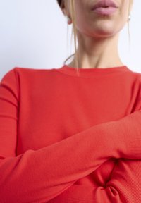 Γυναίκα με κόκκινη μακρυμάνικη μπλούζα και σταυρωμένα τα χέρια, με το πηγούνι και τα χείλη εμφανή, μπροστά σε απλό ανοιχτόχρωμο φόντο.