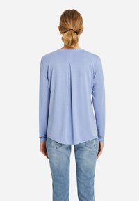 Mujer con cabello rubio recogido en una coleta, vestida con una camiseta de manga larga azul claro y jeans azules, vista desde atrás sobre un fondo blanco.