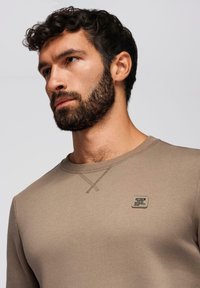 Beige mikina s kulatým výstřihem, která má prošívaný vzor na rameni a malou značenou nášivku na hrudi.