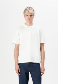 Short-sleeve, cream polo shirt made of soft fabric, featuring a classic collar and two buttons. Paired with dark blue jeans.