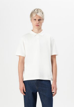 Polo beige à manches courtes en tissu doux, avec un col classique et deux boutons. Associé à un jean bleu foncé.