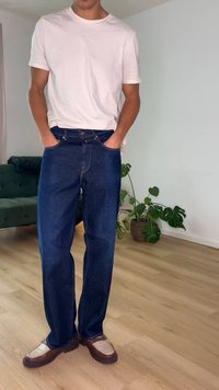 Άνδρας που φοράει απλό λευκό μπλουζάκι, σκούρο μπλε τζιν και καφέ μοκασίνια, στέκεται μέσα σε εσωτερικό χώρο πάνω σε ξύλινο δάπεδο με φυτά και έναν πράσινο καναπέ κοντά.