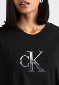 Černé bavlněné tričko s vyšitým stříbrným logem "CK" a textem "Calvin Klein Jeans" níže. Design s kulatým výstřihem a hladkou texturou.
