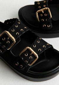 Sandales noires avec deux larges sangles présentant des boucles en or et des œillets. Fabriquées en matériau lisse avec une semelle noire épaisse.