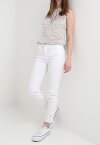 Jean blanc ajusté avec des boutons décoratifs aux poignets, associé à un haut sans manches léger à motifs. Le mannequin porte des baskets blanches.