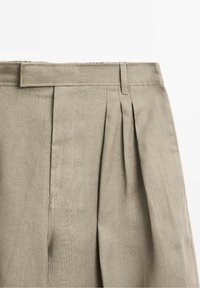 Pantalon en lin beige avec plis devant, ceinture, passants de ceinture et détail sur la couture latérale sur fond blanc.